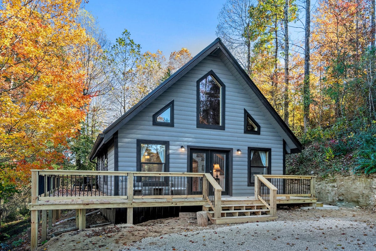 The cabin's exterior showcases modern architecture with large windows framed by seasonal foliage. A spacious deck with a wooden railing extends from the front, providing multiple access points to the entrance. Natural light reflects off the structure, highlighting the inviting surroundings.