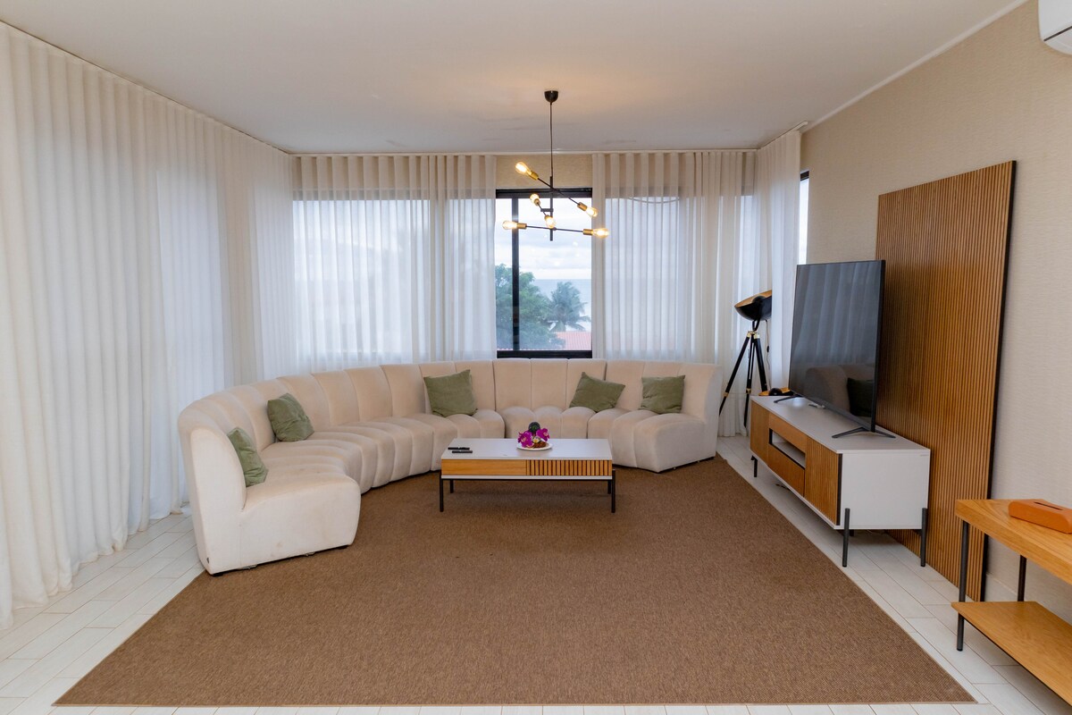 A spacious living area features a curved sectional sofa adorned with decorative cushions. Natural light filters through sheer curtains, illuminating the room. A modern coffee table rests on a textured area rug, complemented by a sleek entertainment unit and a large television.