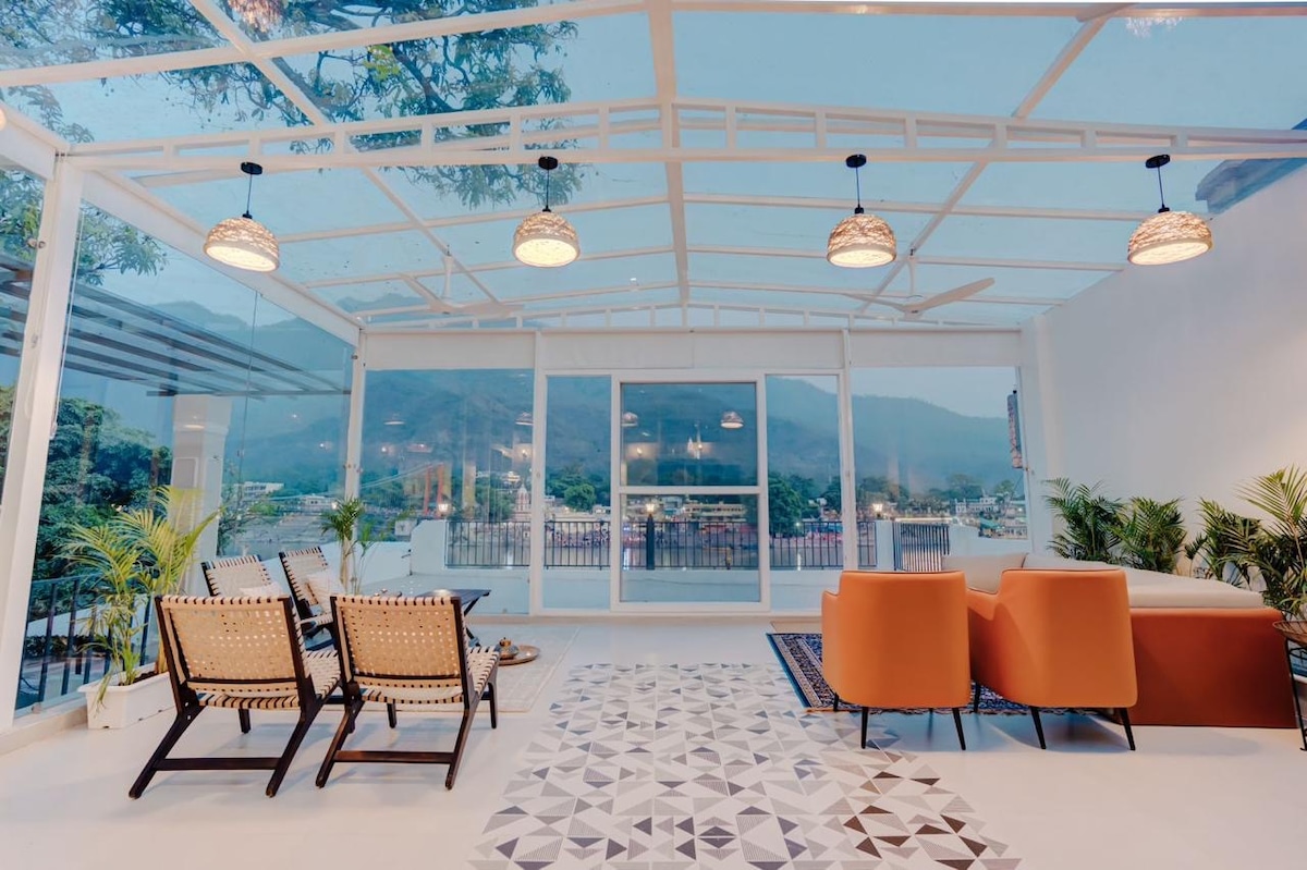 A glass-walled living space offers expansive views of the surrounding mountains. Comfortable seating arrangements include two orange chairs and a patterned rug beneath. Potted plants add a touch of greenery, while stylish light fixtures hang from the ceiling, enhancing the airy atmosphere.