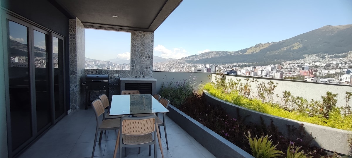 An outdoor terrace is depicted, featuring a glass dining table surrounded by several chairs. A built-in grill is visible nearby, with a backdrop of the city and mountains creating a scenic view.