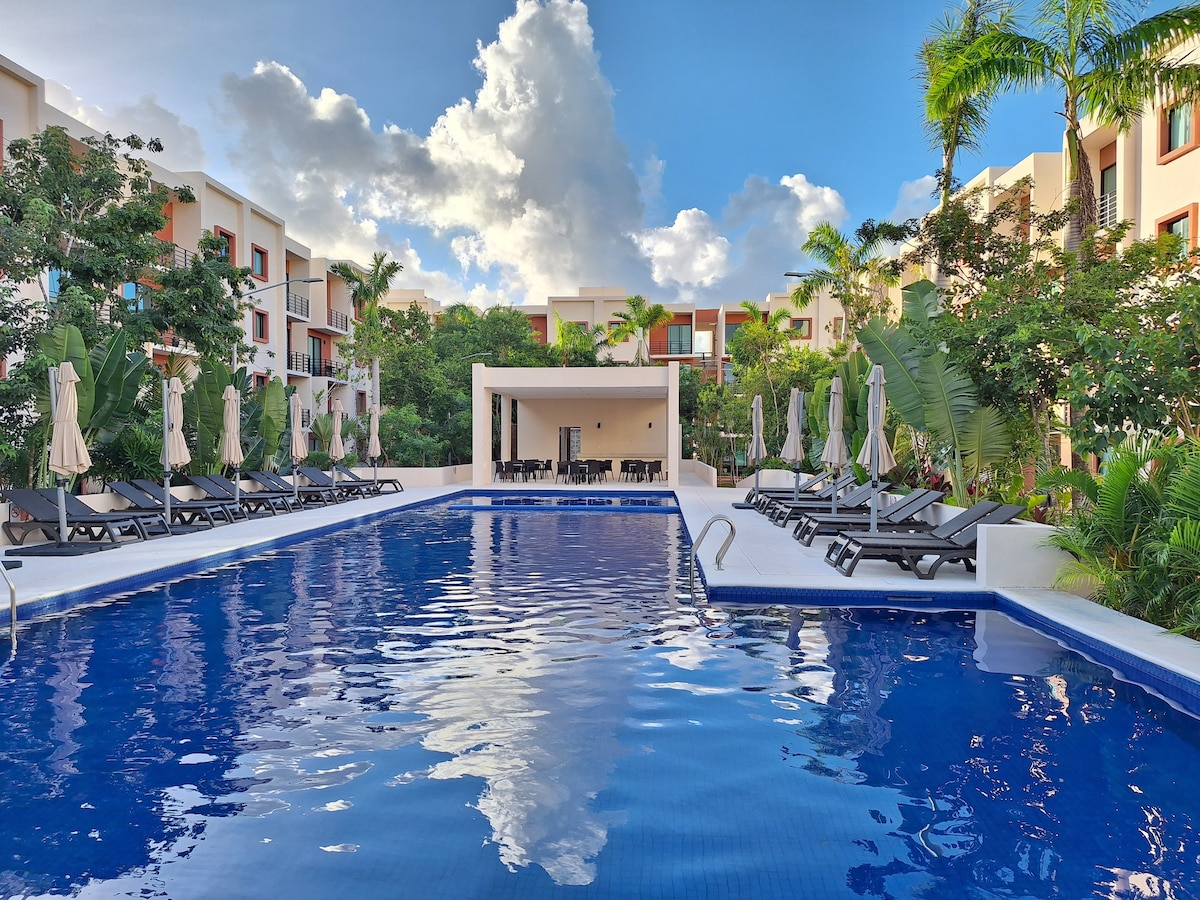 A large swimming pool is surrounded by lounge chairs and umbrellas, with lush landscaping providing a backdrop. The area is designed for relaxation, showcasing vibrant plants and a clear blue sky reflecting in the water. A shaded area with tables is visible nearby.