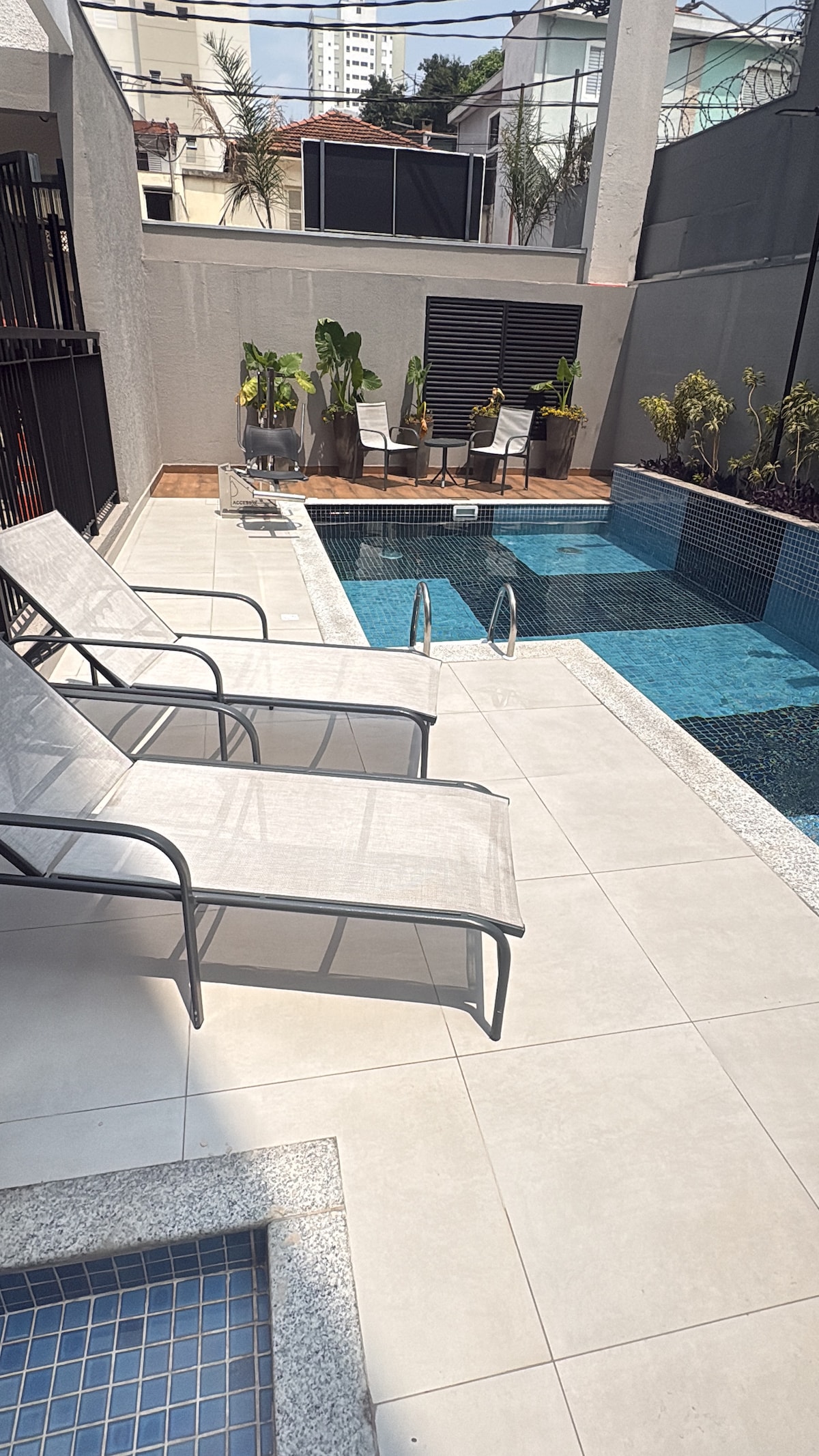 A sunlit area is presented, featuring two lounge chairs positioned beside a sparkling pool. The pool area is surrounded by tropical plants and modern furnishings, creating a serene outdoor space for relaxation.