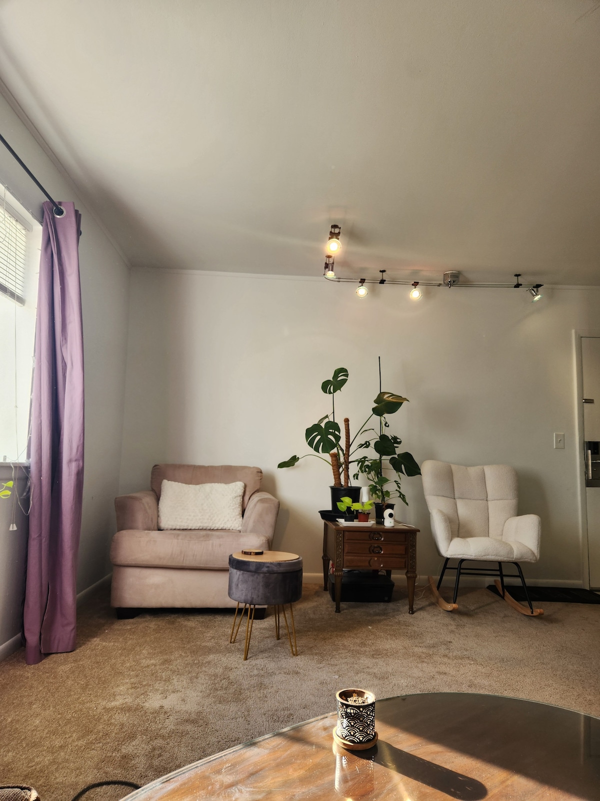 A cozy corner is presented with two comfortable chairs, one upholstered in a soft fabric and the other a modern rocking chair. A small side table sits between them, adorned with a plant. Natural light filters through a nearby window, complemented by light-colored walls and a carpeted floor.