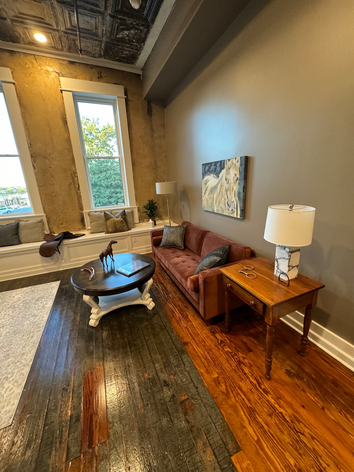 A cozy seating area features a velvety sofa in rich tones, paired with a round coffee table. A lamp with a marble base stands beside a wooden side table, complementing the textured wall and large windows that invite natural light into the space.