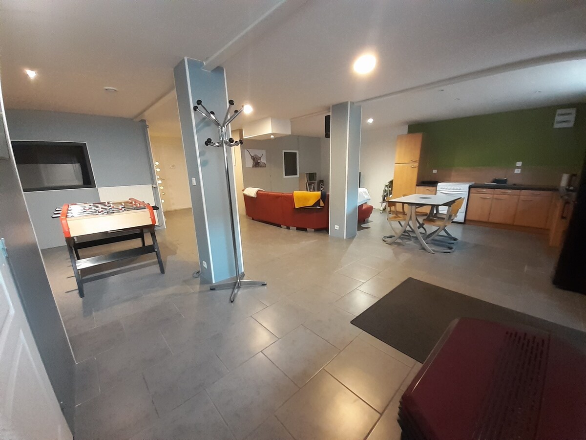 An open-plan living space features a foosball table beside a coat rack. A sectional sofa and a dining area with a folding table are visible. The kitchen is equipped with wooden cabinets, and natural light enters through a window, illuminating the polished floor.