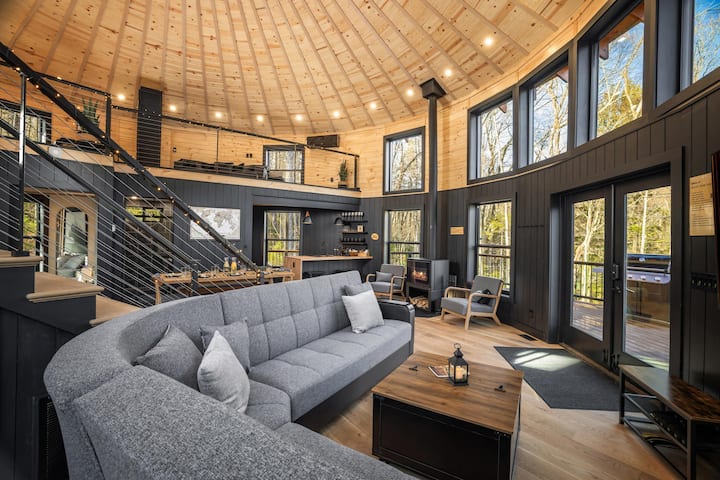 Comfortable sectional in the open living area beside the loft staircase and wood stove, with forest views through wraparound windows.