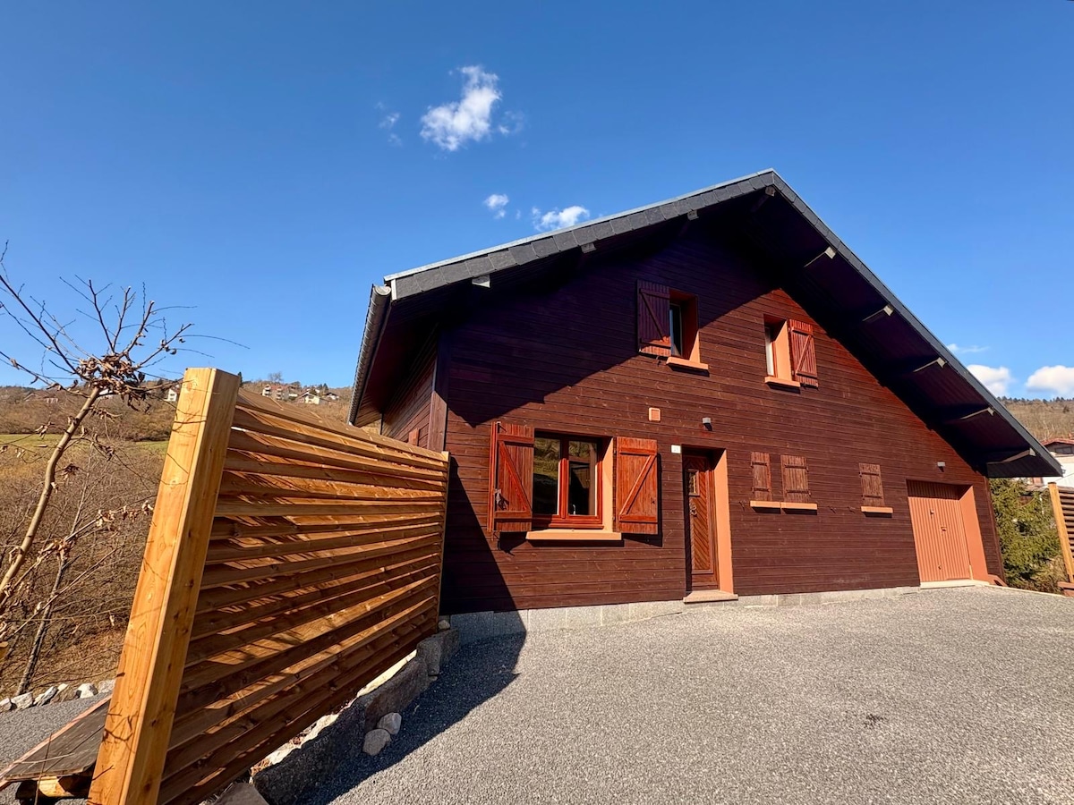 A modern wooden chalet is shown against a clear blue sky, featuring gabled rooflines and large windows framed by wooden shutters. The exterior is complemented by a gravel driveway and a wooden fence, highlighting the natural surroundings.