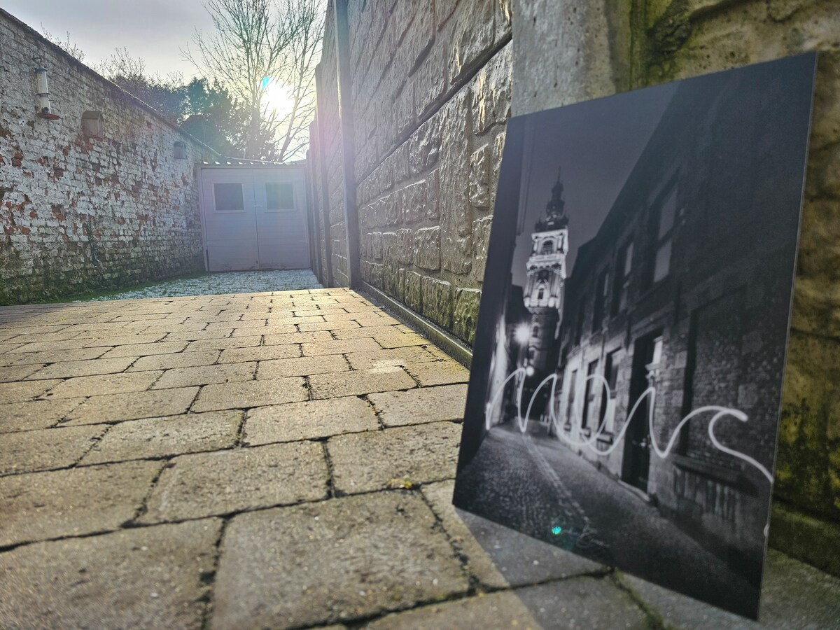 A narrow cobblestone alley is depicted, leading toward a distant building illuminated by soft sunlight. A black and white photograph rests on the ground, featuring a historical structure adorned with lights. Textural variations of the stone walls frame the scene.