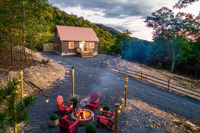A charming log cabin sits nestled among trees, featuring a welcoming front porch. A circular fire pit surrounded by red Adirondack chairs provides a cozy gathering space. Soft light illuminates the scene, highlighting the natural surroundings and nearby gravel pathway.