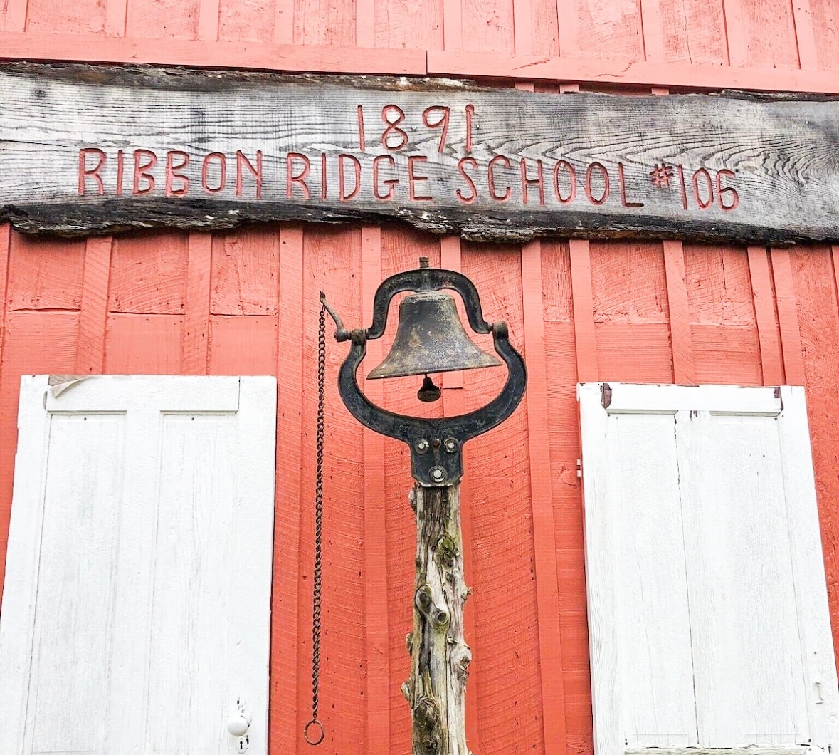 The exterior of the Ribbon Ridge Schoolhouse is showcased with a bright red wooden structure. Above the entrance, the year '1891' is inscribed on a rustic sign. A bell is mounted on a sturdy post, complementing the vintage aesthetic of the property.