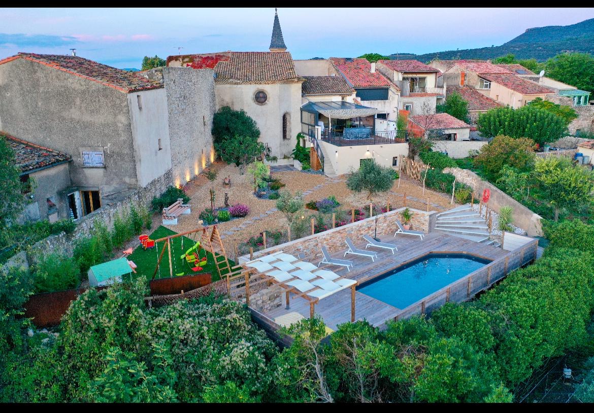 An aerial view captures the spacious garden, featuring a private pool surrounded by lounge chairs. A children's play area with swings and a slide is visible, along with a mix of stone paths and landscaped greenery. The unique chapel adds character to the picturesque setting.