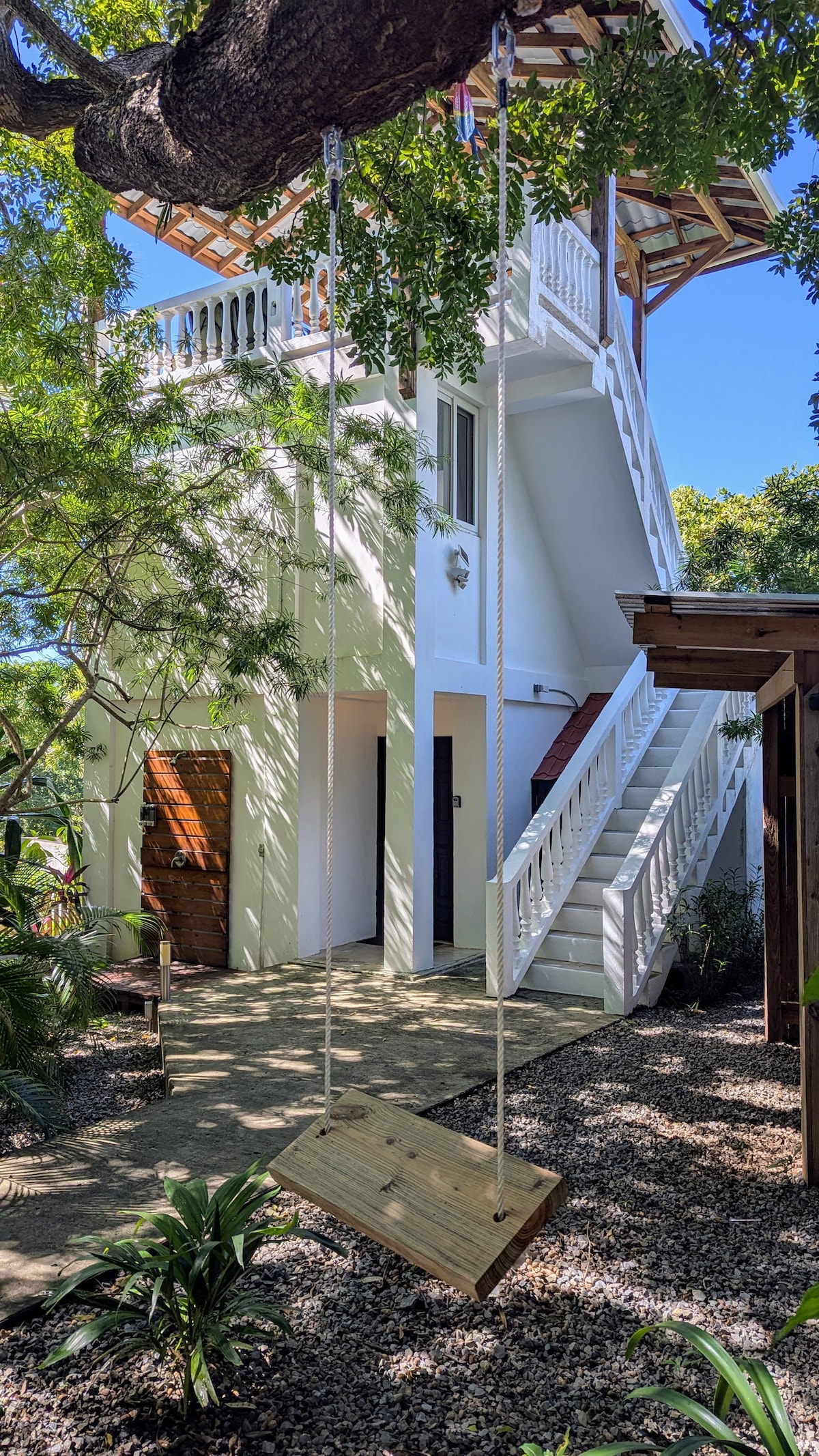 A wooden swing hangs from a tree branch in a shaded garden area, offering a tranquil spot for relaxation. The white building in the background features a staircase leading to the upper level, surrounded by lush greenery and gravel pathways.