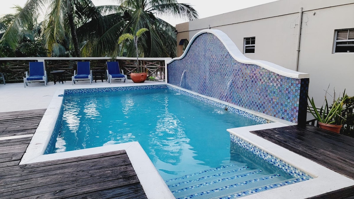 A swimming pool with vibrant mosaic tiles is set within a spacious outdoor area. Lounge chairs are arranged nearby, and lush palm trees provide a tropical backdrop. The pool features a waterfall edge, enhancing the inviting atmosphere of the space.