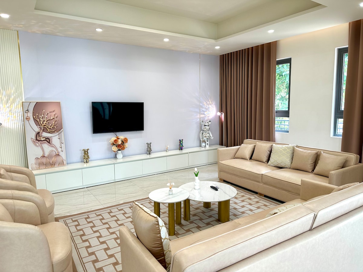 A spacious living area features a light-colored sectional sofa and four individual chairs arranged around a round coffee table. A large flat-screen TV is mounted on a white wall, accompanied by decorative accents and a light decor palette. Natural light filters through the windows with soft curtains.