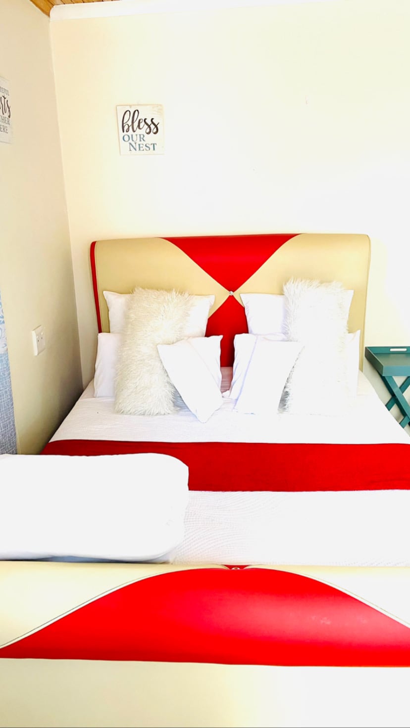 A bed with a red and cream headboard features an arrangement of multiple white pillows, creating a comfortable sleeping area. A neatly folded white towel is placed at the foot of the bed. A green side table stands beside the bed, contributing to the overall decor.