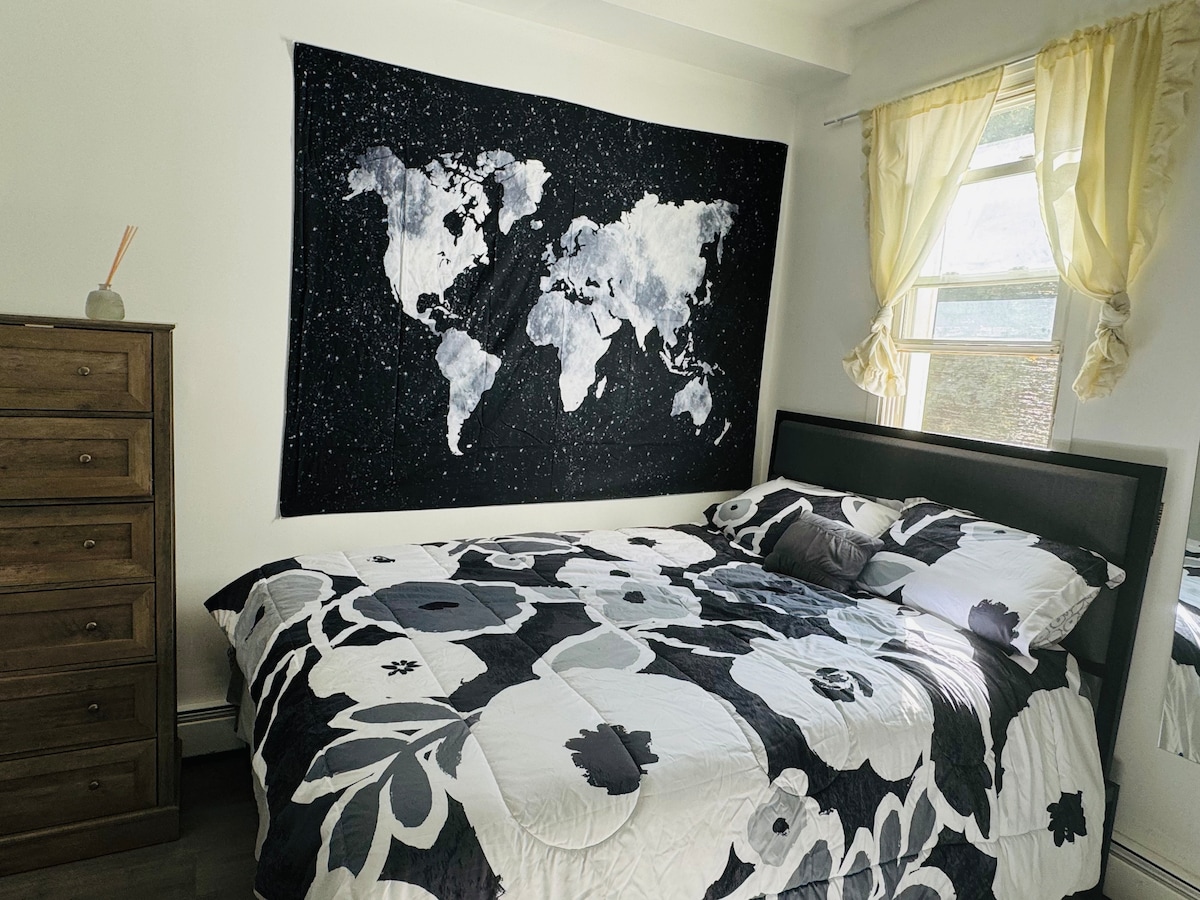 A cozy bedroom features a large bed covered with a floral-patterned quilt. A tapestry displaying a world map adorns the wall above the bed. Natural light enters through a window draped with light curtains, and a wooden dresser is positioned nearby.