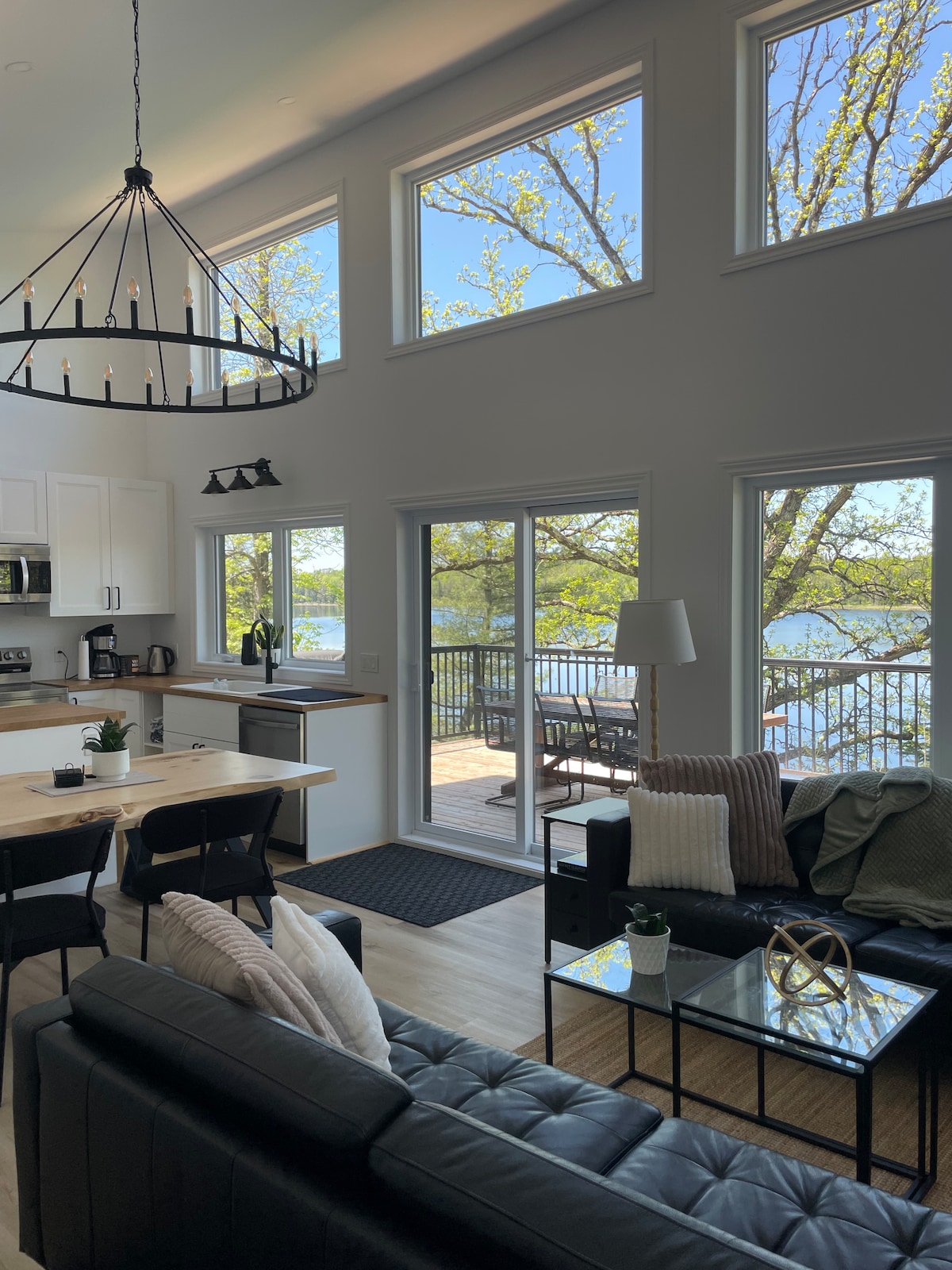 A modern living space is showcased with large windows that invite natural light and offer views of the lake. A comfortable black sofa is positioned beside a glass coffee table, while a dining table accommodates four. The kitchen area features sleek cabinetry and appliances.