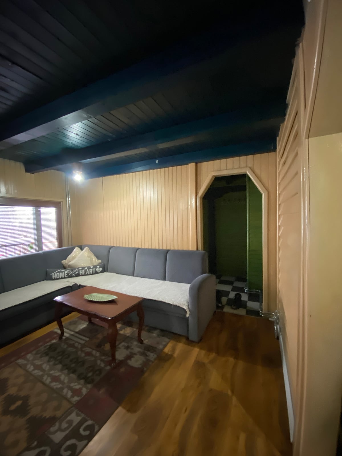 A seating area features a comfortable gray sofa with decorative pillows and a wooden coffee table at its center. Light enters through windows, illuminating the wooden floor and textured walls. An arched doorway leads to a softly lit space, enhancing the room's cozy ambiance.