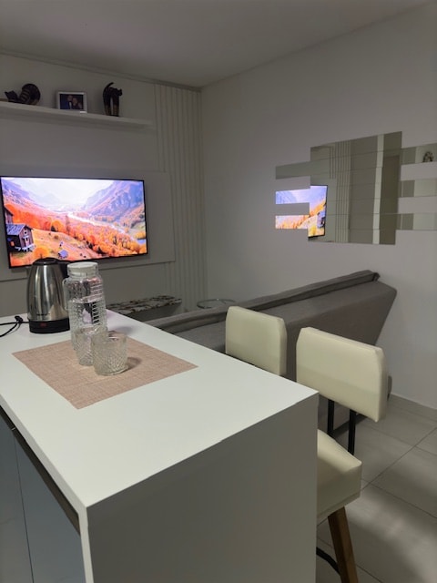 A modern living area is visible with a sleek white countertop, accompanied by two tall white chairs. A television mounted on the wall displays a scenic mountain view. A gray sofa is positioned against the wall, with a decorative mirror reflecting the light.