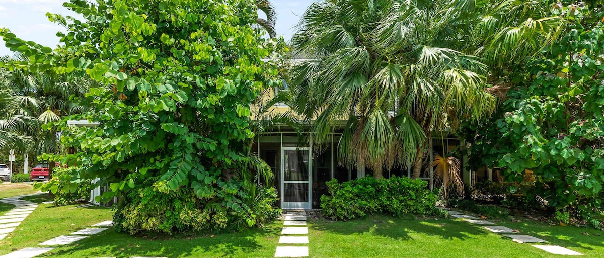 The entrance features a screened front door framed by lush greenery, including tall palm trees and vibrant plants. A well-maintained pathway of stepping stones leads through the manicured lawn, welcoming guests into the serene surroundings.