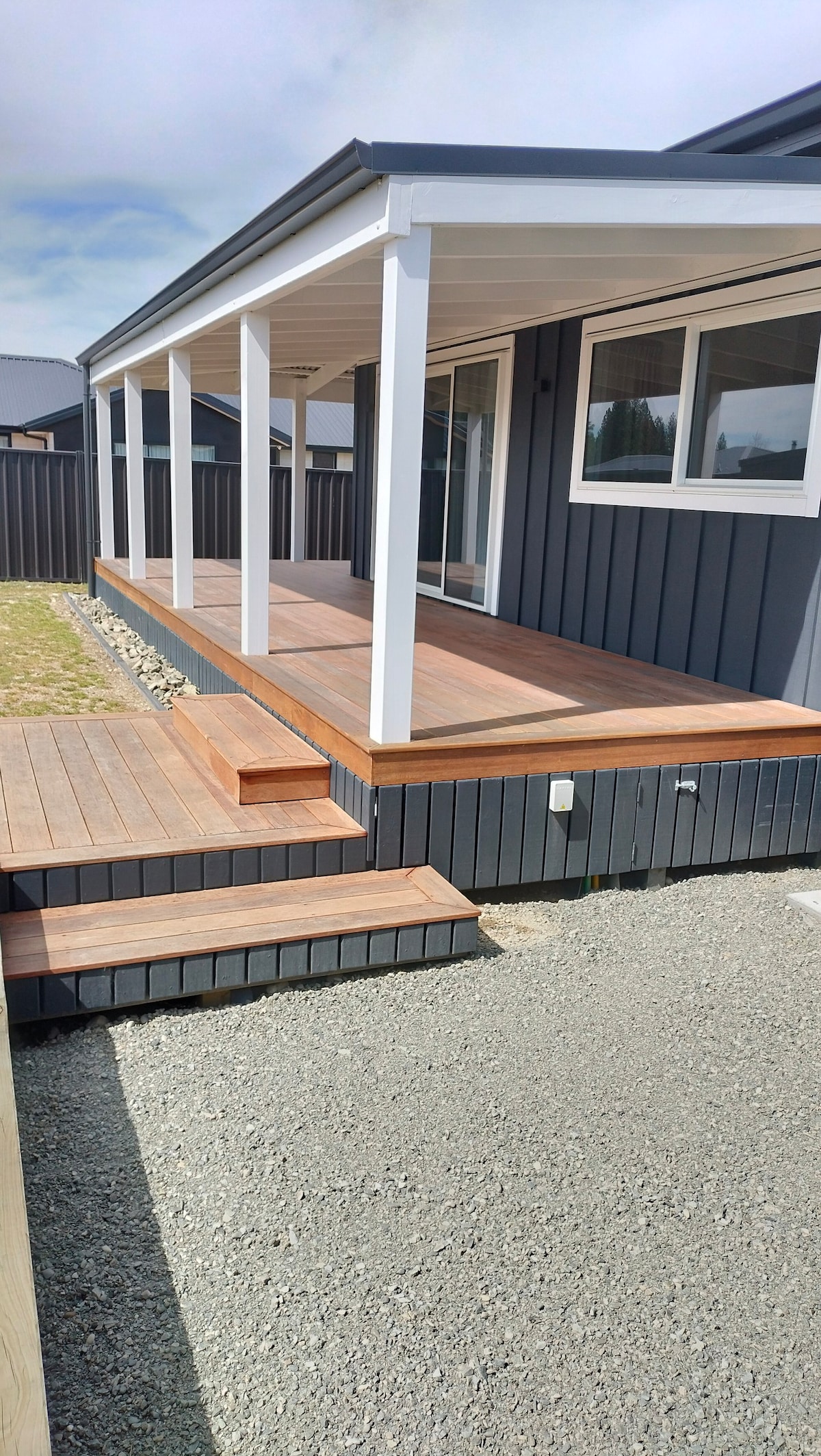 A covered veranda is featured, accessed by a set of wooden steps. The grey exterior of the cottage contrasts with the warm-toned decking. Light enters through large windows, enhancing the inviting outdoor space.