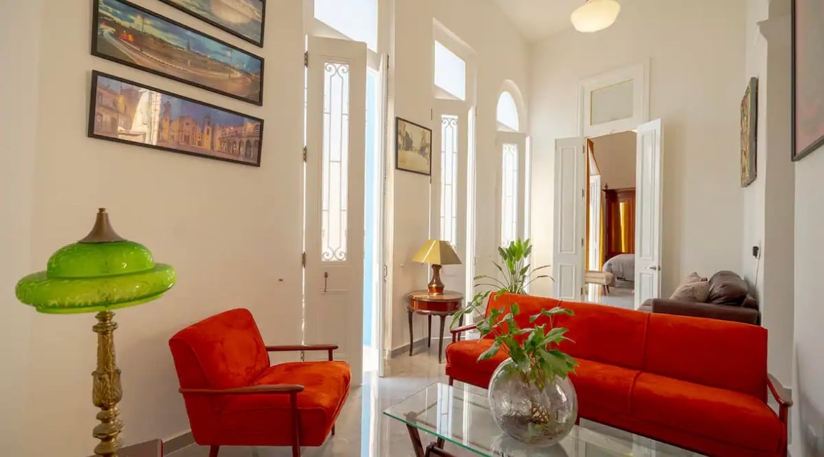 A bright living area features large windows allowing natural light to flow in. An orange sofa is paired with a matching armchair, while a glass coffee table sits in the center. Decorative art pieces adorn the walls, contributing to the warm ambiance.