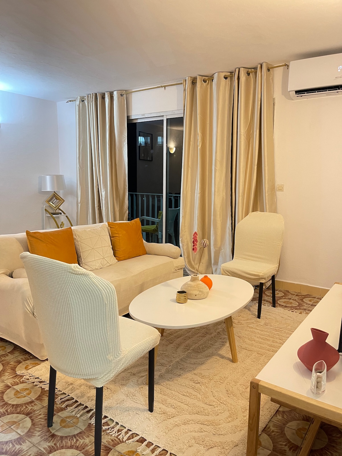 A cozy living area is presented with a light-colored sofa adorned with decorative cushions. Two chairs flank a circular coffee table, accompanied by a small vase and a decorative object. Natural light filters through sheer curtains, enhancing the room's relaxed ambiance.