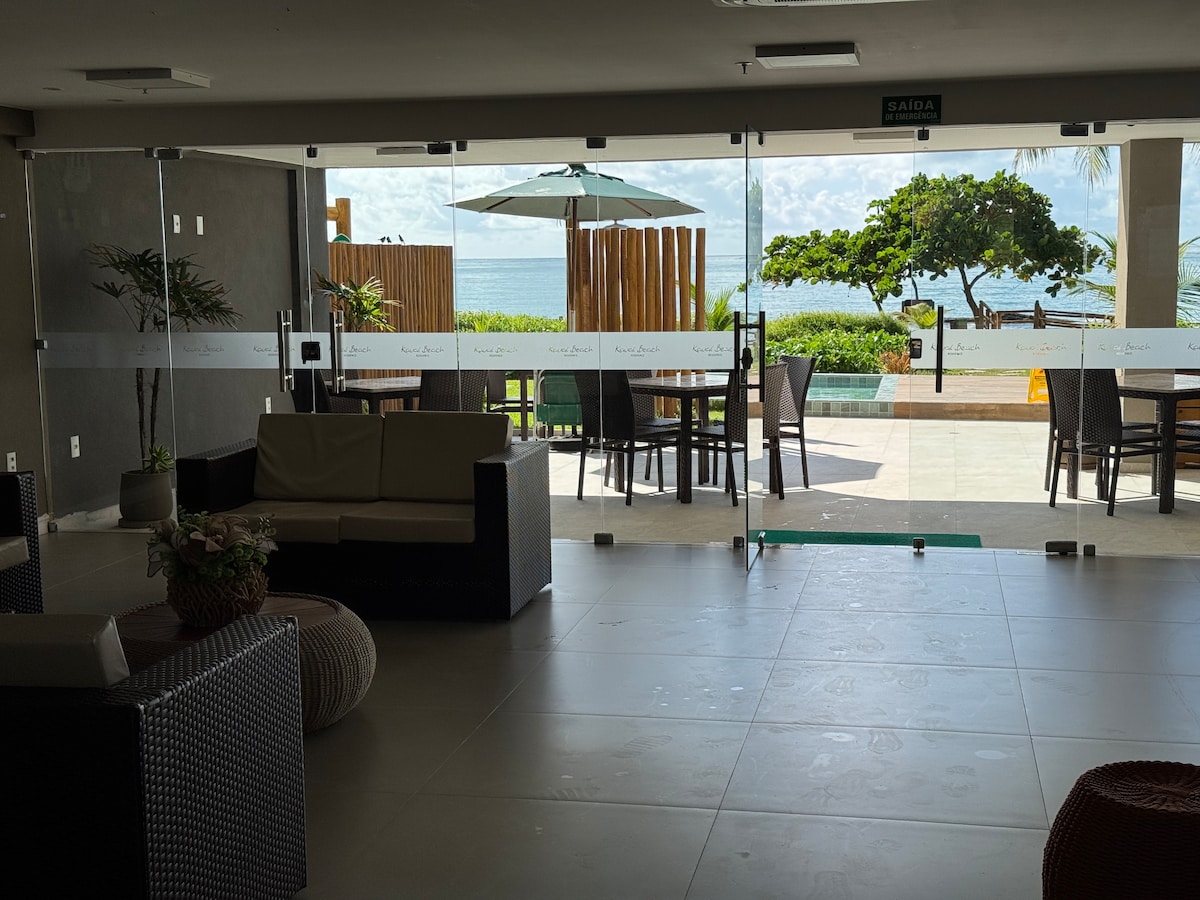 The image captures a spacious lobby area with modern furnishings. Large glass doors provide a clear view of the outdoor patio, featuring an umbrella over a table and ocean views. Comfortable seating arrangements are positioned within the lobby, enhancing the inviting atmosphere.