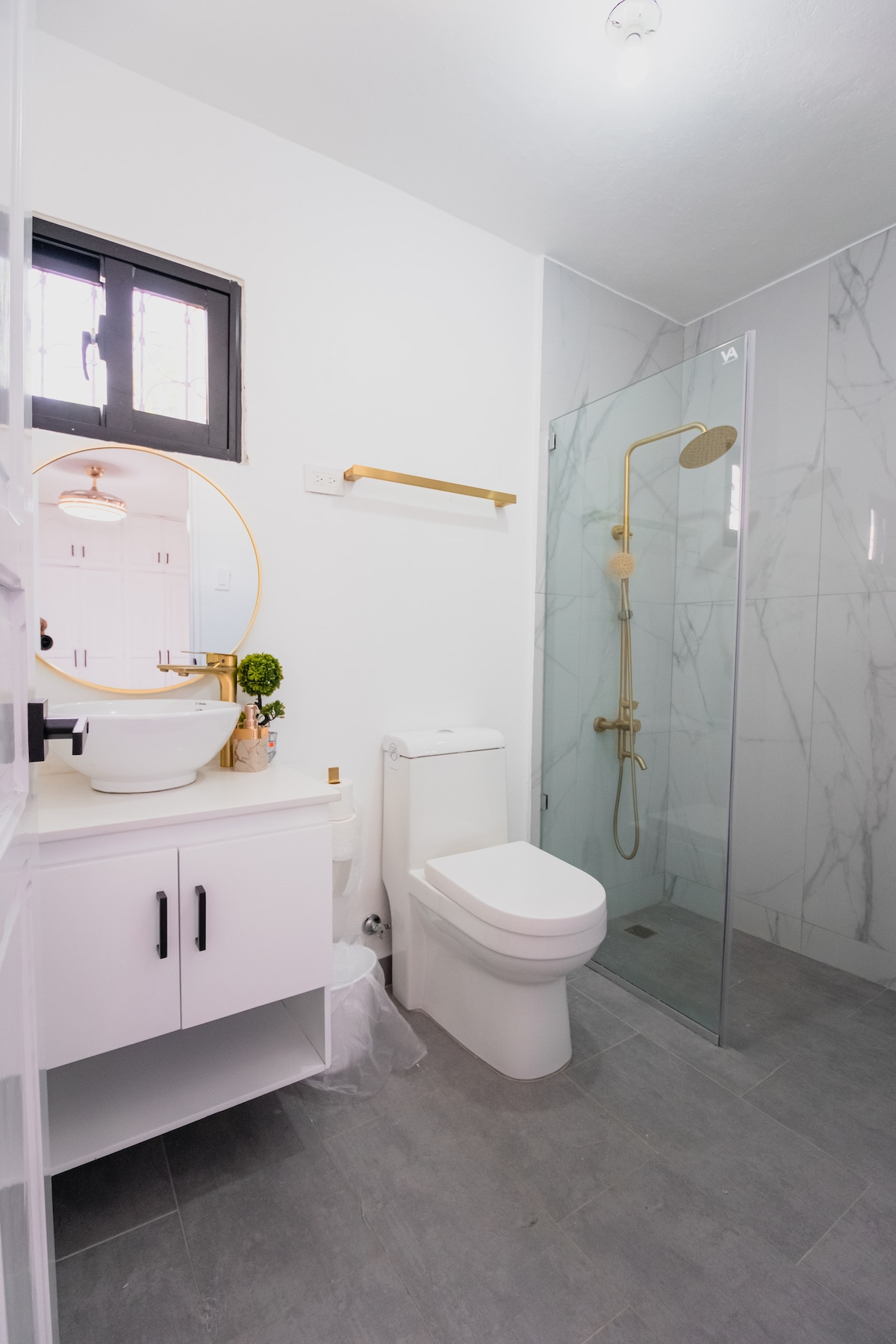 A modern bathroom is displayed with elegant marble accents on the walls. A circular mirror is mounted above a sleek vanity with storage. A glass shower enclosure features a gold rainfall showerhead, complemented by minimalist decor and neutral tones throughout.