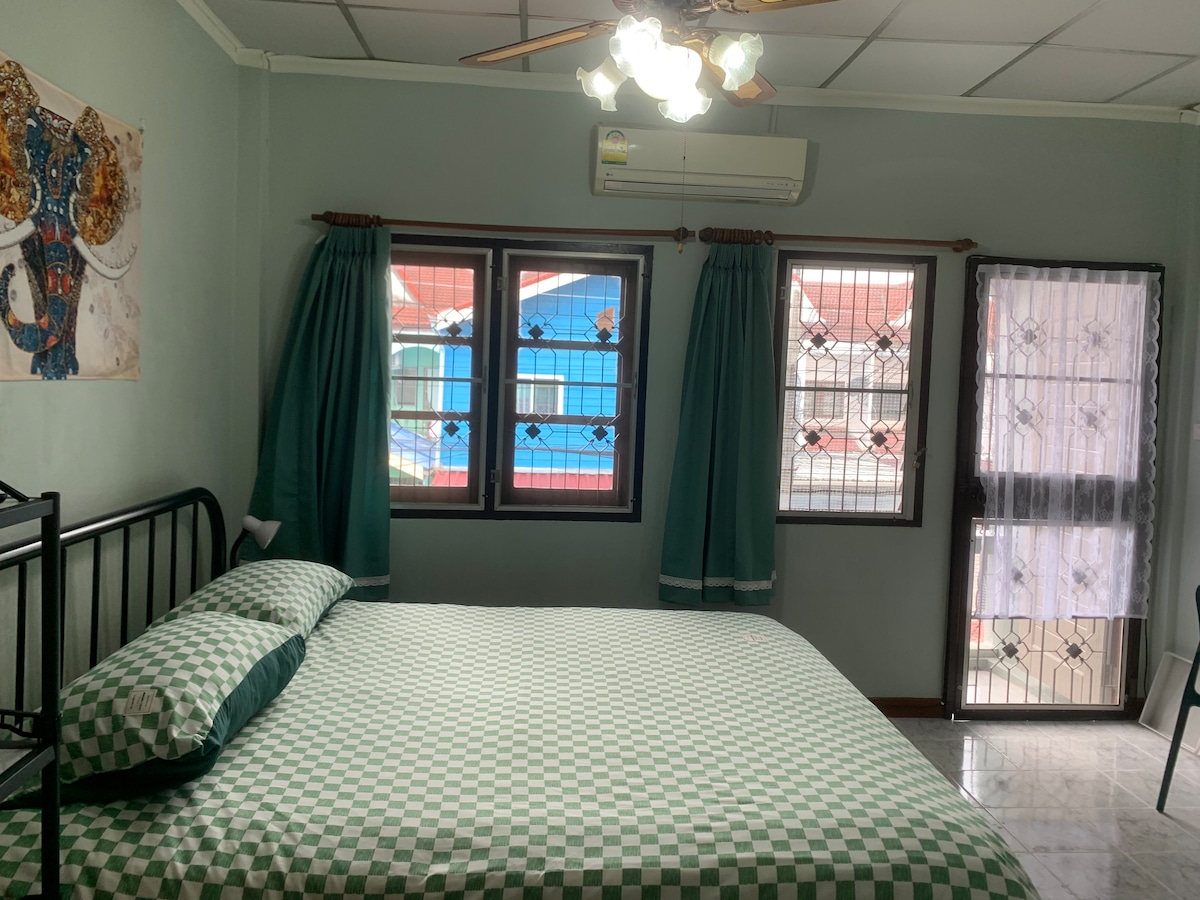 A spacious room features a large bed with a checkered green and white duvet. Two windows are dressed with green curtains, allowing natural light to fill the space. A ceiling fan and air conditioning unit provide comfort. An elephant-themed artwork is displayed on the wall.
