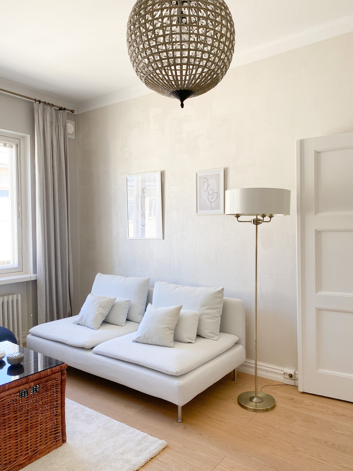 A bright living area features a simple white sofa adorned with several light blue accent pillows. A floor lamp stands nearby next to a wicker table. Large windows allow ample natural light to illuminate the space, and soft, neutral curtains frame the windows.