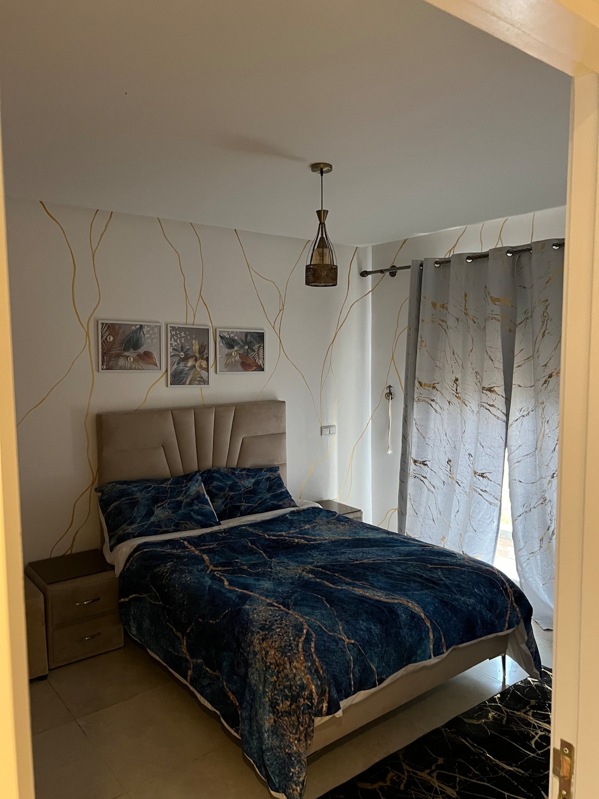 A compact bedroom features a plush bed with a blue and gold patterned bedspread. Two framed artworks hang above the headboard. Natural light filters through sheer, patterned curtains, and a small bedside table is positioned next to the bed, enhancing the cozy environment.