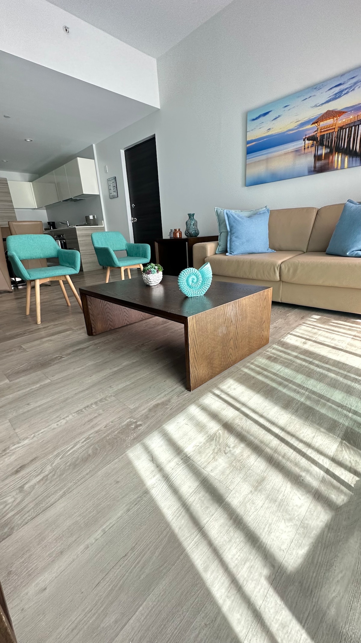 A modern living space is highlighted, featuring a beige sofa accompanied by two turquoise chairs. A wooden coffee table is centered, complemented by decorative items. Natural light casts soft shadows on the floor, while a beach-themed wall art piece adds a tranquil touch.