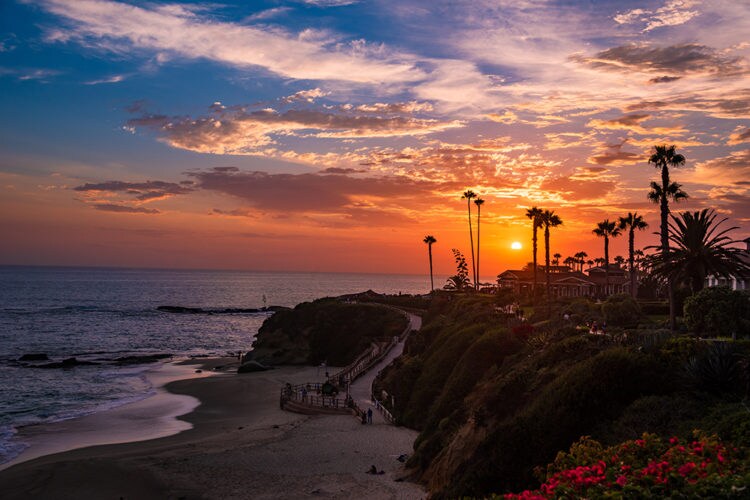 A serene beach scene captures a vibrant sunset casting warm hues over the ocean. Silhouetted palm trees line the coast, framing the view of the sandy shoreline. Gentle waves lap at the beach, while a pathway winds along the landscape, inviting peaceful exploration.