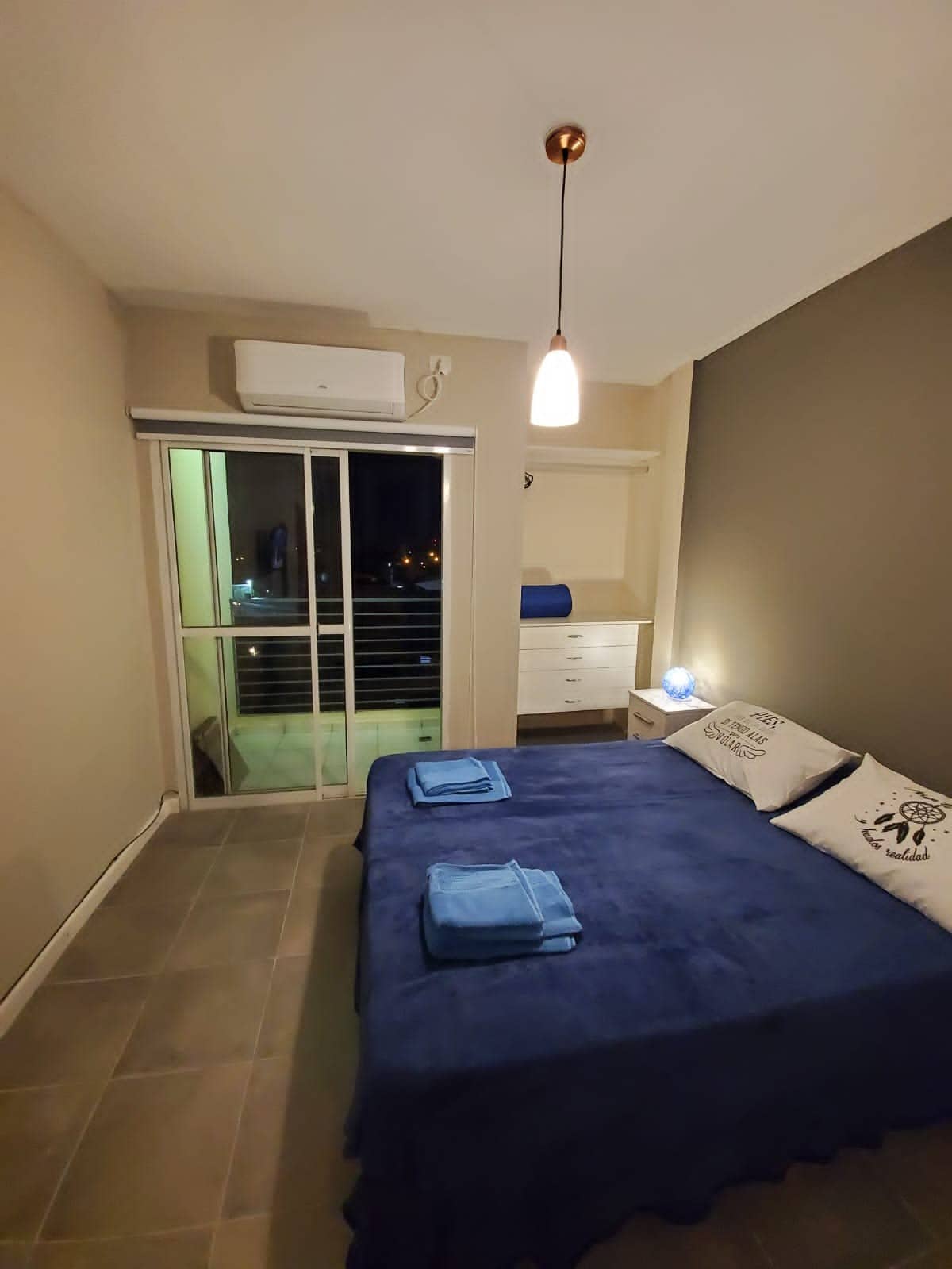 A modern bedroom features a spacious bed covered with a blue blanket. A sliding door leads to a small balcony, providing a view of the surroundings. A light fixture hangs from the ceiling, and a dresser is positioned against the wall.