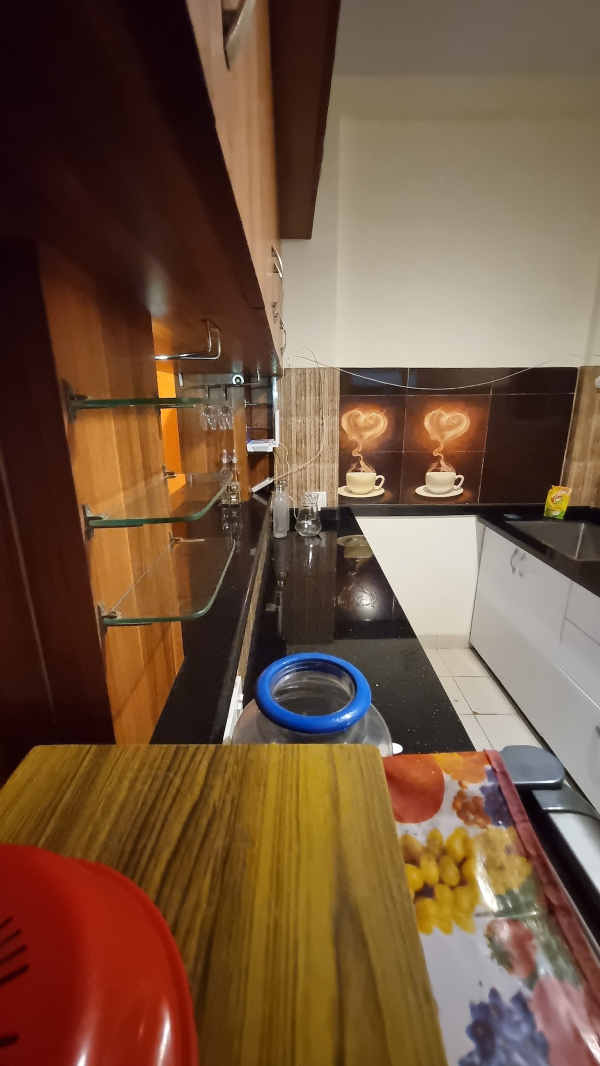 The kitchen features a sleek black countertop combined with wooden cabinetry. Open shelves are positioned along the wall, displaying a few decorative items. The backsplash showcases an artistic design, while the modern appliances stand ready for use.