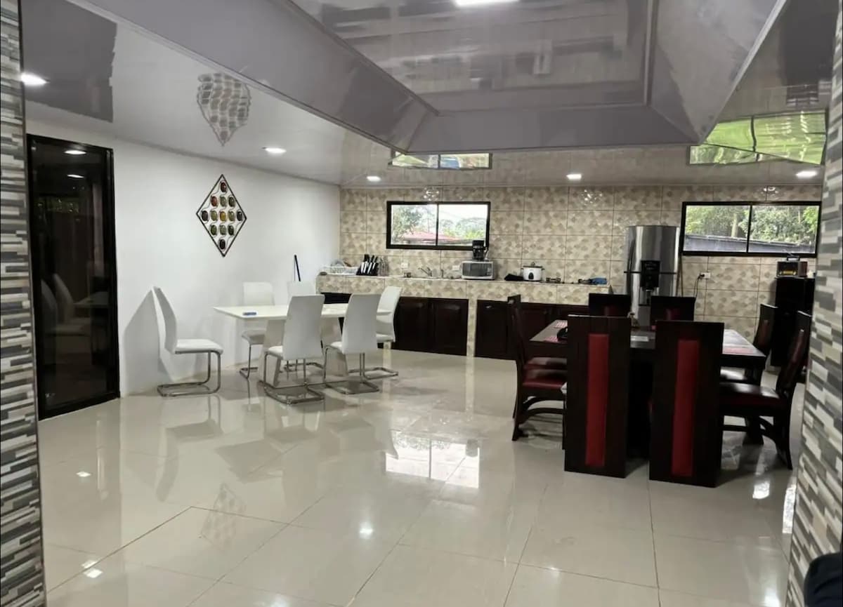 A modern dining area is presented, featuring a mix of white and dark wooden furniture. Six white chairs surround a rectangular table, while an additional set of four dark chairs and a large table is visible. Bright, reflective tile floors contribute to the room's spacious ambiance.