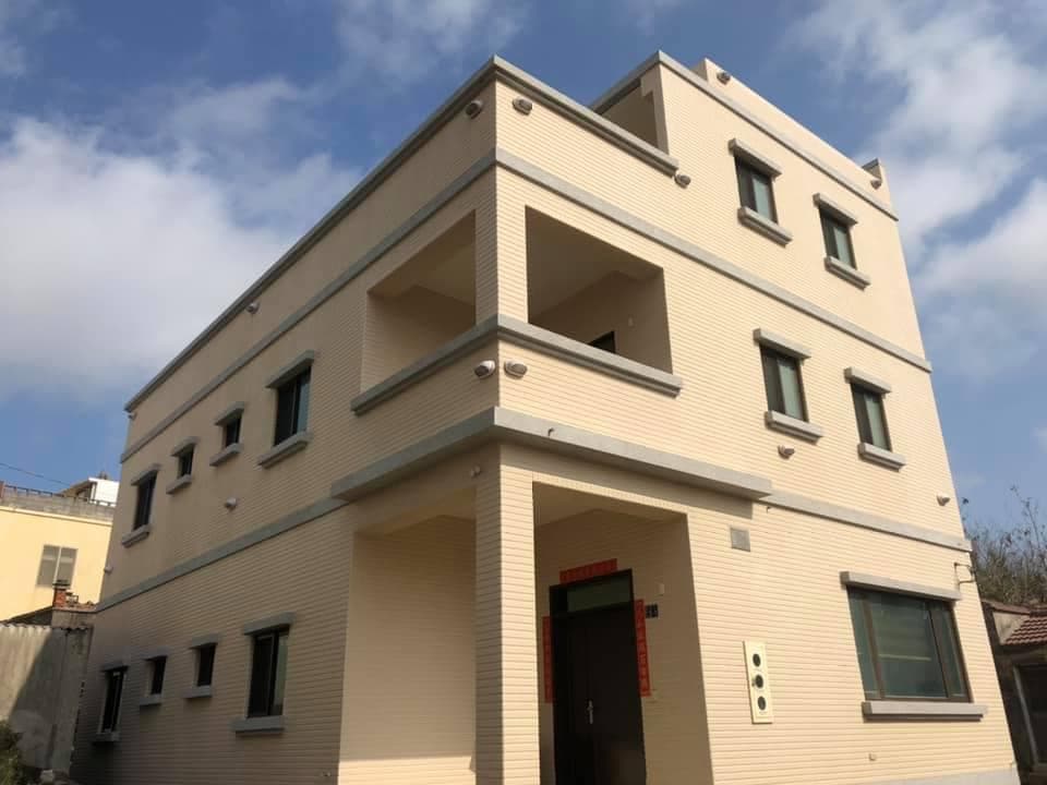 A three-story building features a light-colored brick façade with multiple windows and balconies. The design incorporates clean lines and a modern aesthetic. A welcoming entrance is visible on the ground level, framed by two strategically placed security lights.