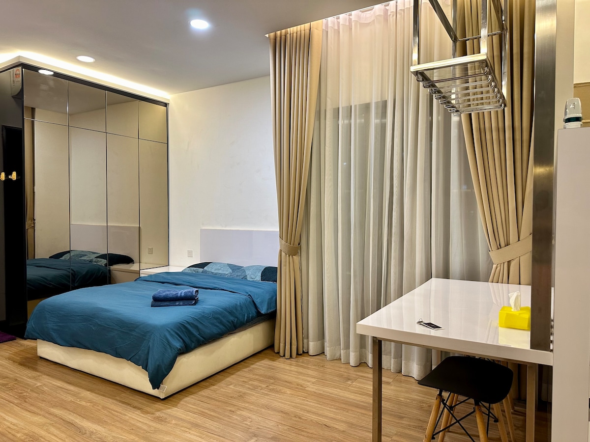 A cozy studio features a spacious bed with a deep blue coverlet. A mirrored wardrobe reflects the soft lighting, while large curtains frame a window. A minimalist desk with a chair is positioned to the side, contributing to the room's functionality.