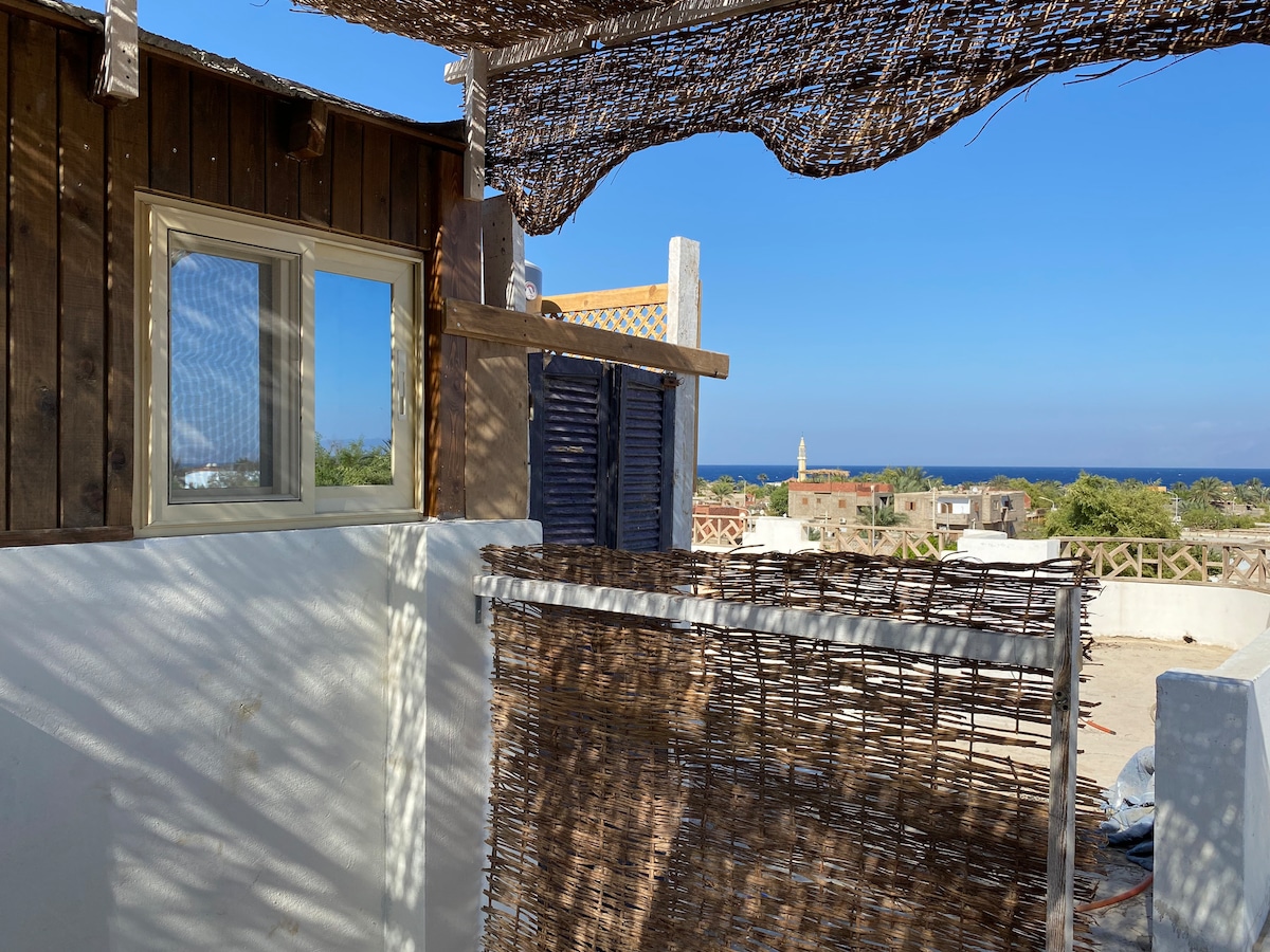 A private rooftop area is shown with rustic wooden panels and woven shading, offering views of the surrounding landscape. A large window reflects natural light, framing the spacious outdoor space against a backdrop of the ocean and clear blue sky.