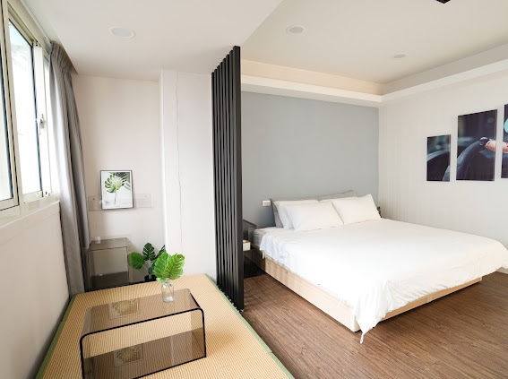 A spacious room features a queen size bed with crisp white bedding, separated by a decorative partition. A low table sits atop a tatami mat, while large windows allow natural light to fill the space. Minimalist decor enhances the serene atmosphere.