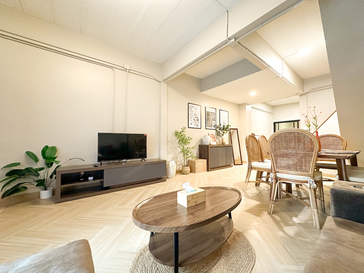 A spacious living area is furnished with a comfortable sofa and a low coffee table. A television is mounted on a modern cabinet, while a dining table with rattan chairs is positioned nearby. Green plants add a touch of nature to the light-filled space.