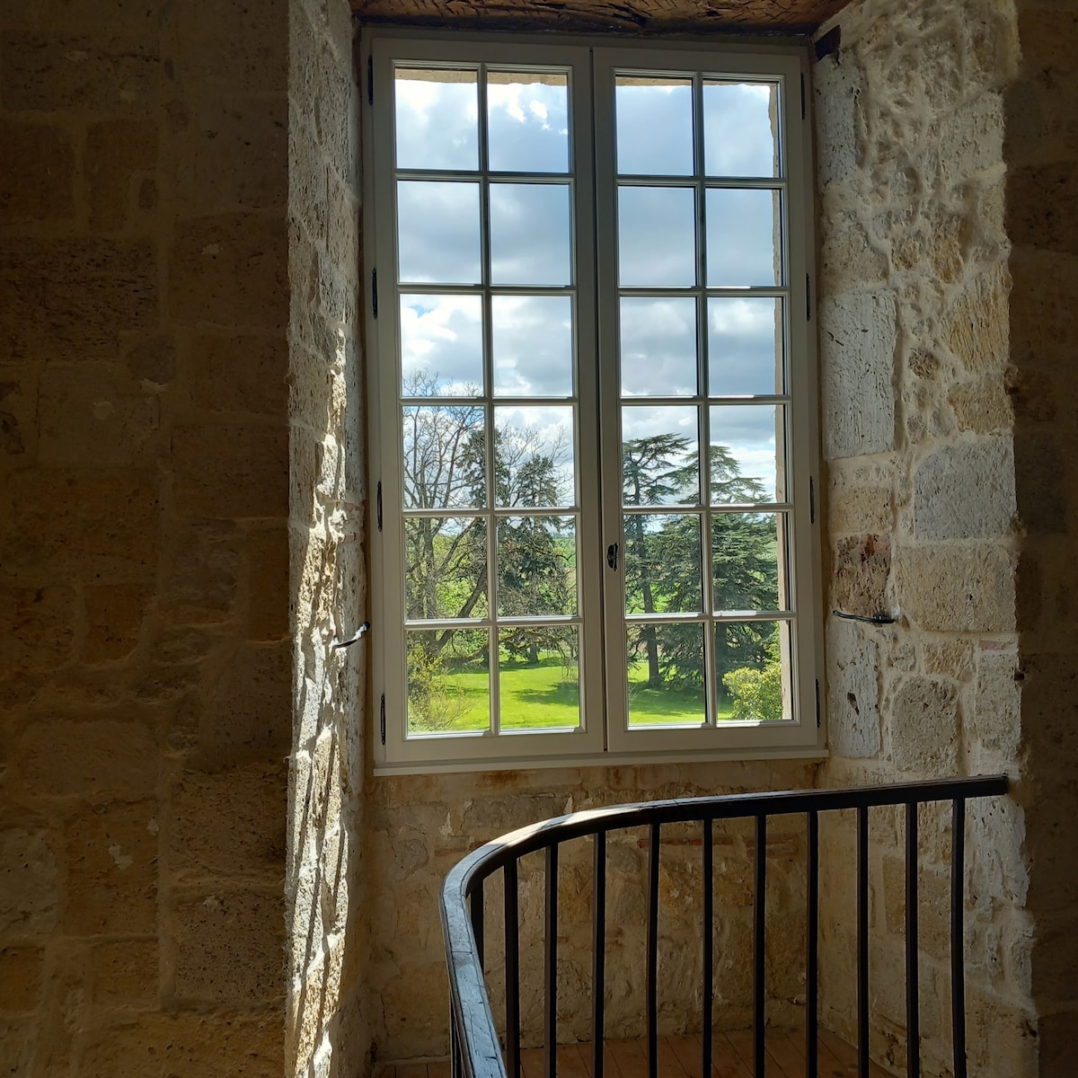 A large window reveals a view of the surrounding greenery and trees outside. Sunlight filters through the glass panes, illuminating the stone interior of the tower. A wrought-iron railing is visible in the foreground, enhancing the architectural character of the space.