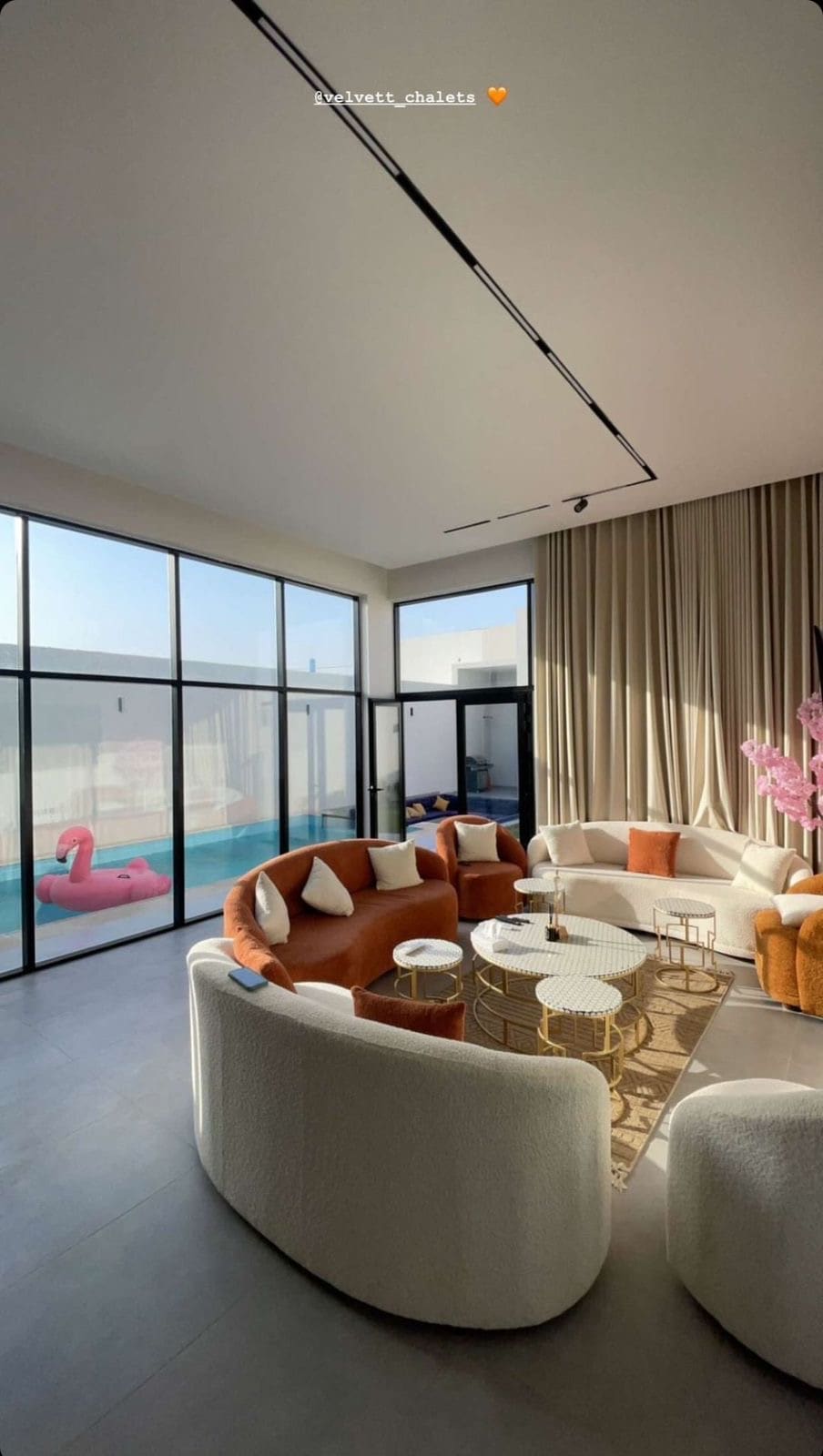 A bright living area is filled with natural light from large glass windows. Soft seating with a mix of neutral and warm tones surrounds a circular coffee table, creating a relaxed atmosphere. A hint of a pool is visible through the windows, along with a pink inflatable float.