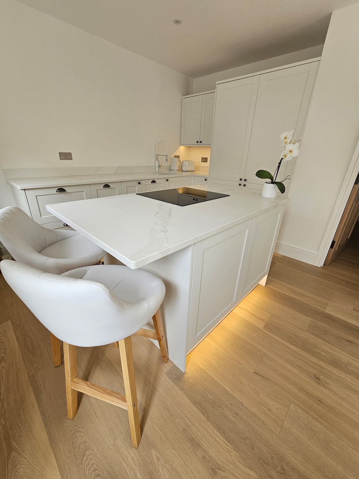 A modern kitchen is highlighted by sleek, white cabinetry and a bright, airy aesthetic. Bar stools with light upholstery are positioned at a stylish island featuring a built-in cooktop. Natural wood flooring enhances the space, illuminated by soft lighting beneath the cabinetry.