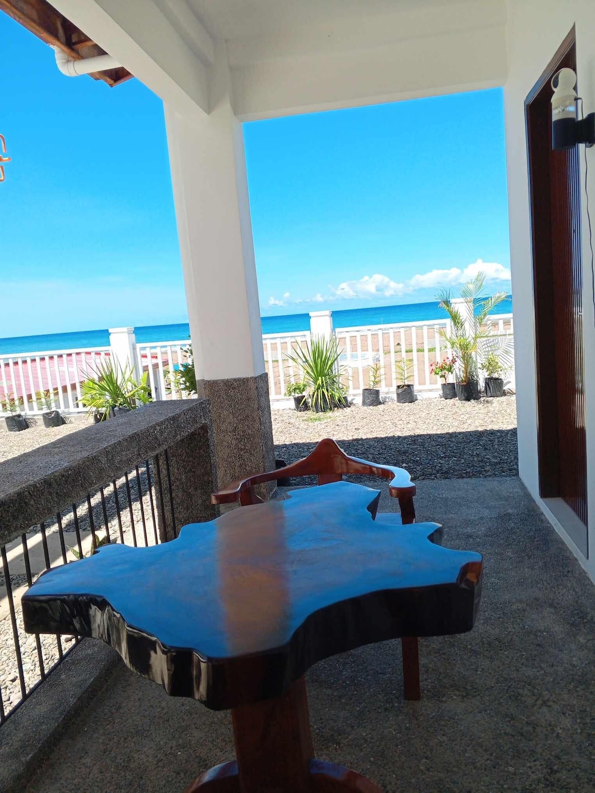 A wooden table with a natural edge design is positioned on a covered veranda, providing a view of the ocean. Lush potted plants line the perimeter, while the bright blue sky complements the serene atmosphere. The scene is framed by white railings and concrete flooring.