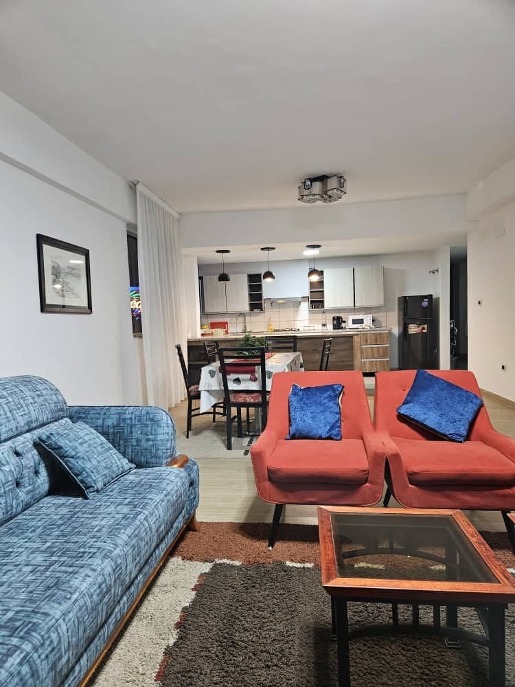 A living area showcases a blue fabric sofa paired with two red armchairs, each adorned with blue cushions. A glass coffee table sits centrally on a textured rug. The open kitchen is visible in the background, featuring modern cabinetry and appliances.
