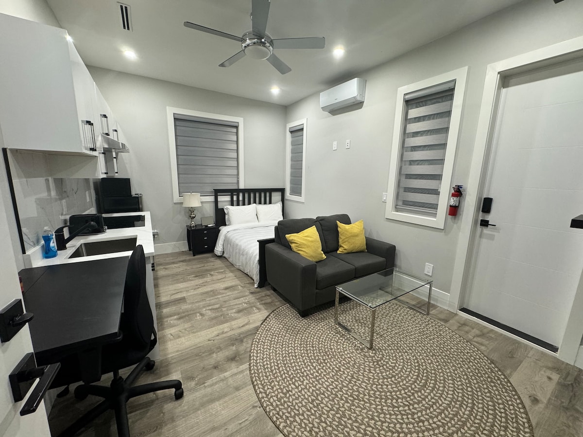 A thoughtfully designed living space features a spacious bed with soft linens and contrasting yellow accent pillows. A modern seating area with a dark gray couch is positioned across from a glass-topped coffee table. A workspace with a black desk and chair is included, and natural light is provided through the window treatments.