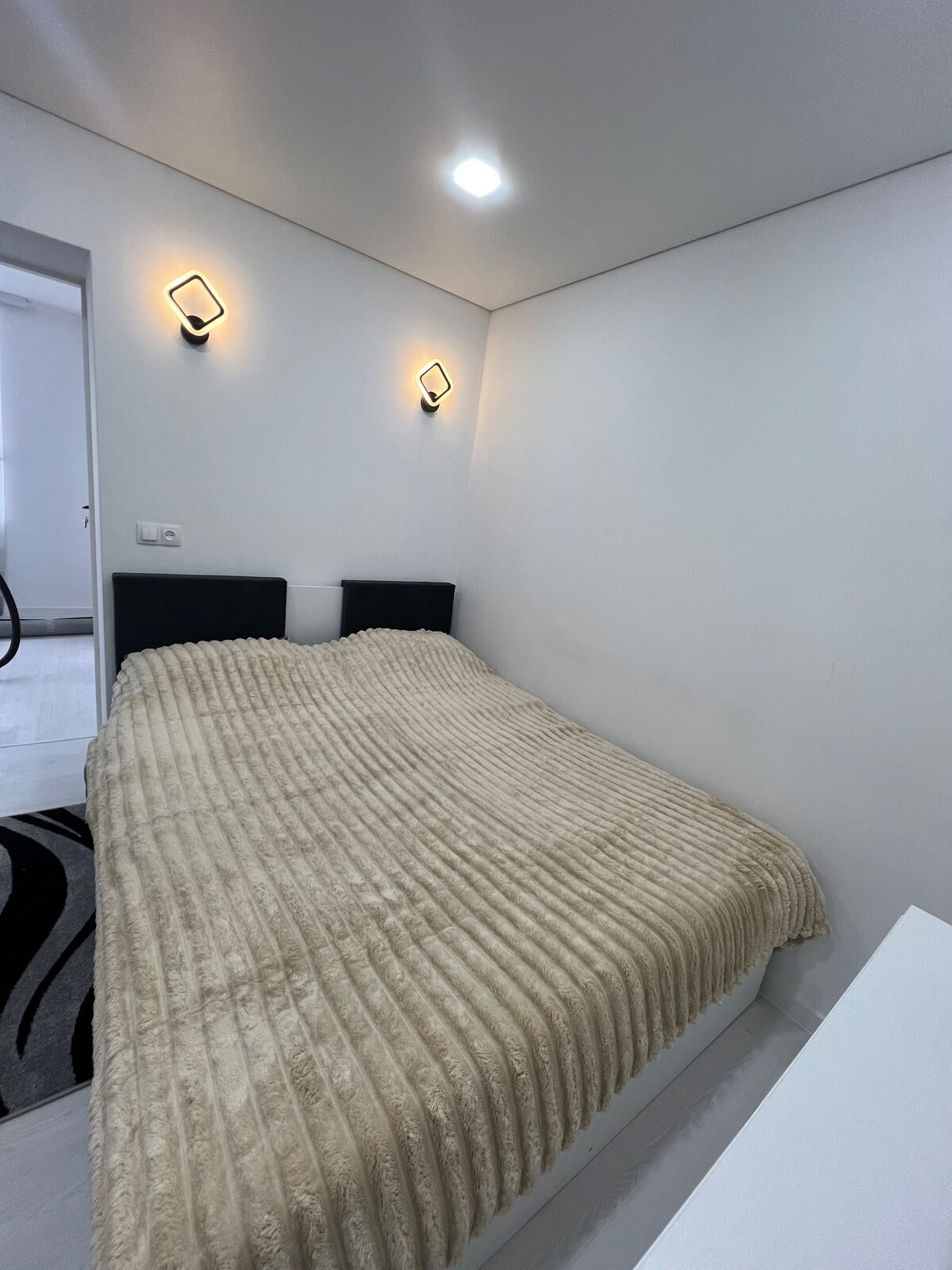 A cozy sleeping area is furnished with a spacious bed draped in a textured cream-colored blanket. Two wall-mounted lamps provide soft lighting, complementing the neutral white walls and the minimalist decor.