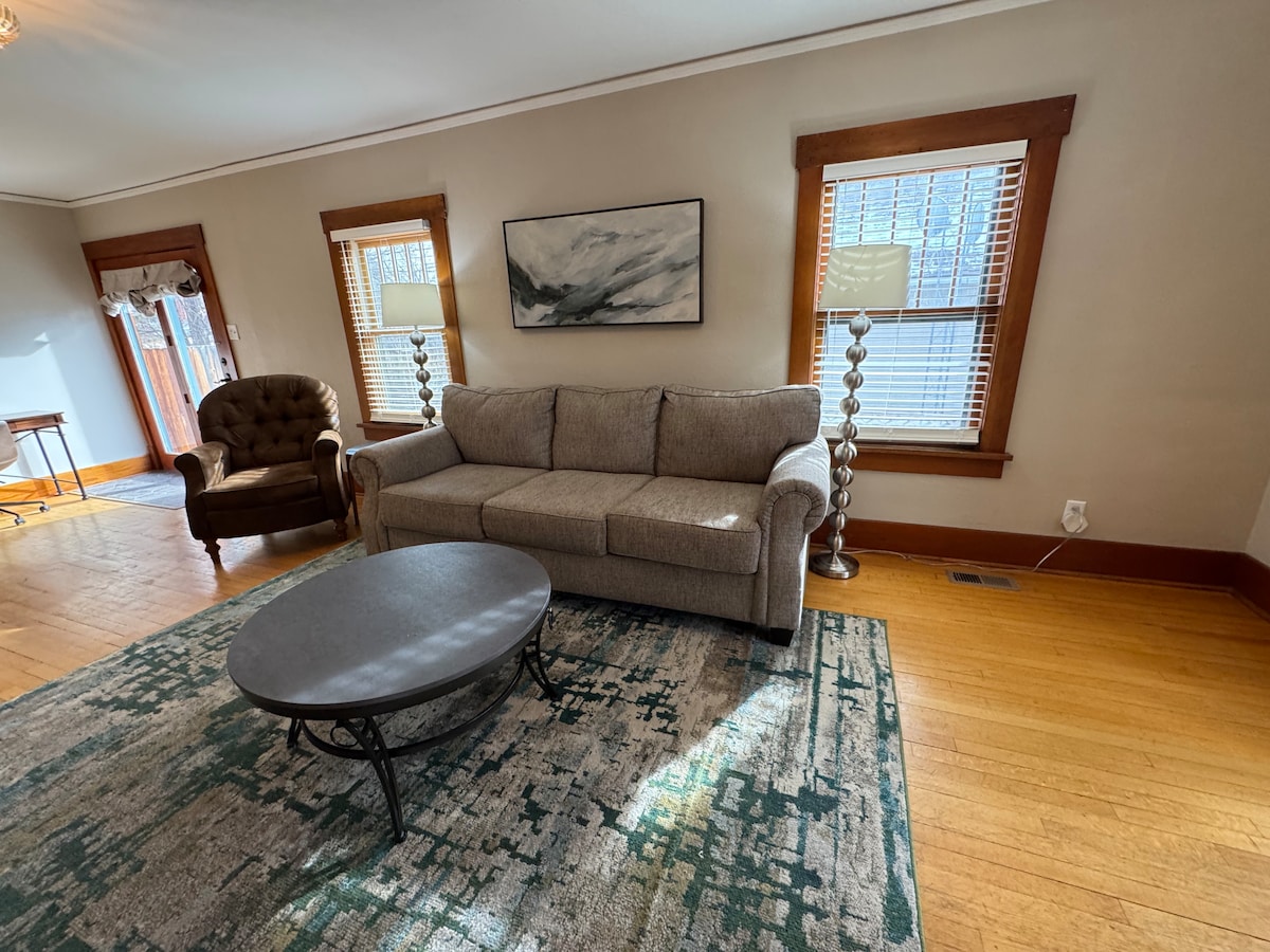 A comfortable living area is showcased, featuring a gray sofa positioned near two large windows with wooden frames. A round coffee table rests on a textured area rug, while a lamp stands beside the sofa, providing additional lighting. A brown armchair adds warmth to the space.